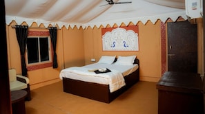 A single-bed room with a ceiling fan, a window with curtains, a decorative headboard, and a small table. - The Desert Resort (Bhuj)
