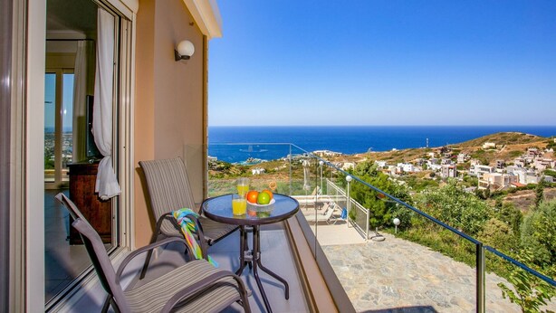 A balcony with a table set for two, a chair, and a view of the sea and town.