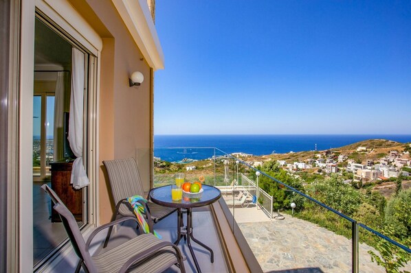 A balcony with a table set for two, a chair, and a view of the sea and town.