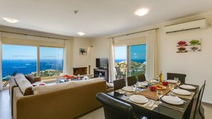 A modern living room with a sofa, dining table set for four, and a TV. Large windows offer a view of the sea and buildings.