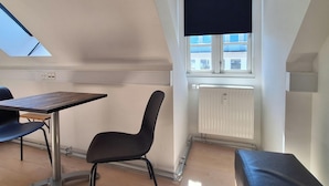 Una habitación moderna y bien iluminada con techo inclinado, una mesa con una silla, un banco y una ventana con persiana.