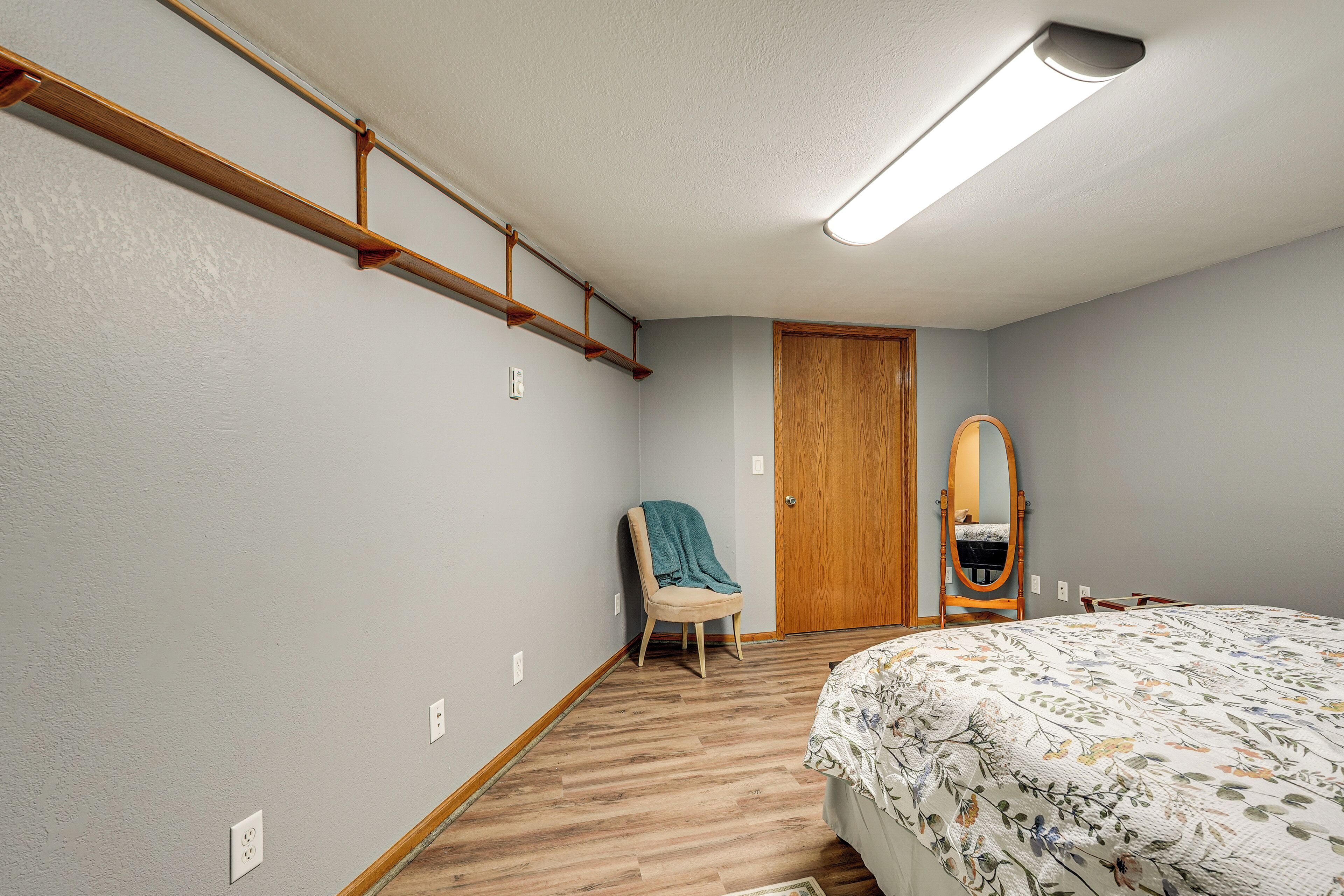 Une chambre avec un plancher en bois, un lit recouvert d’une courtepointe à motifs floraux, une chaise avec une couverture, une porte en bois et un miroir accroché au mur.