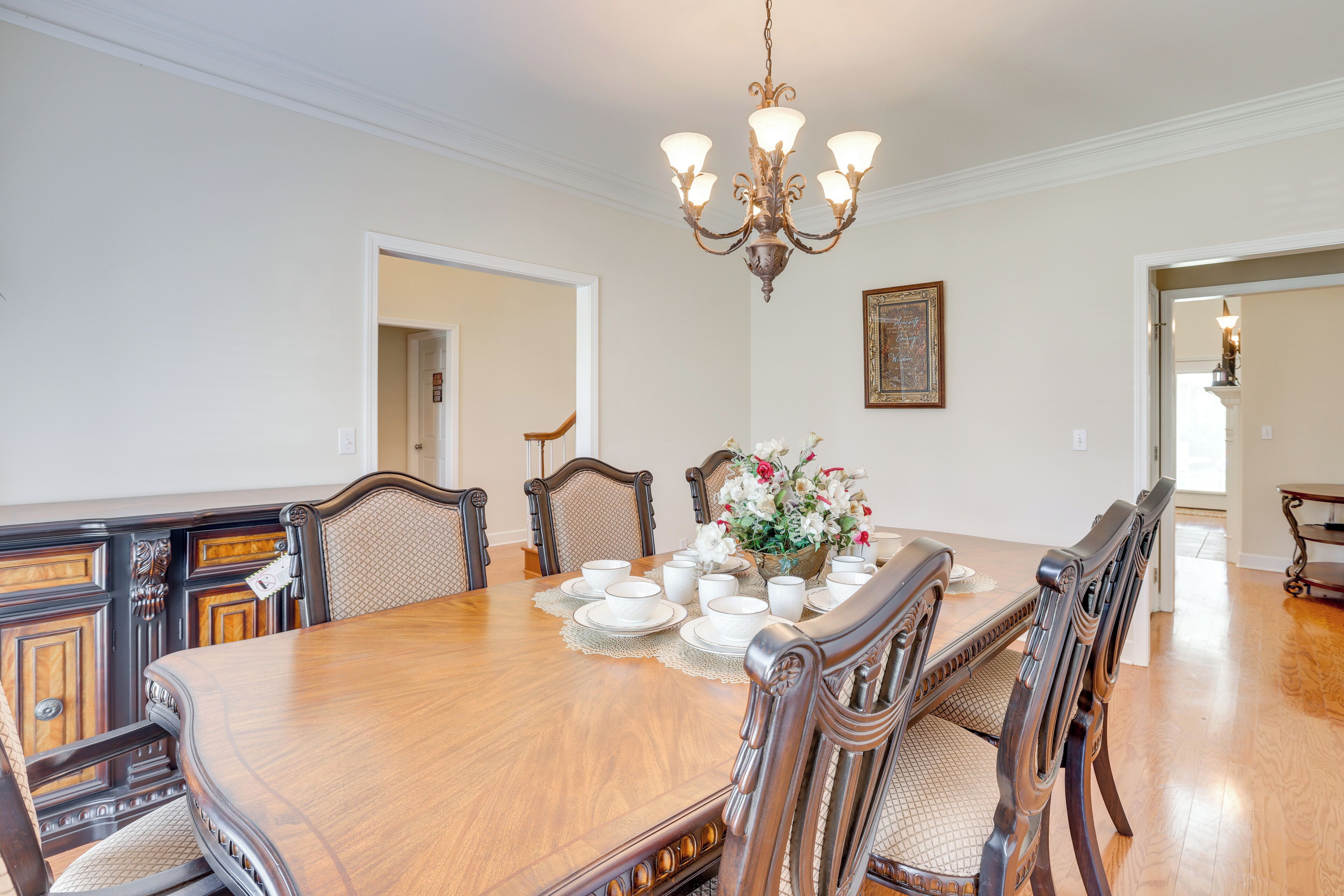 A dining room with a large wooden table, six chairs, a chandelier, and a vase with flowers.