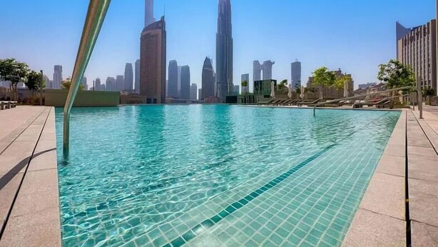 Ein Schwimmbecken mit klarem, blauem Wasser, umgeben von einer gefliesten Terrasse und einer Stadtansicht im Hintergrund.
