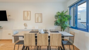 A modern dining area with a long table, four chairs, two wall-mounted artworks, a potted plant, and a window with a view. - Holiday Home 'Oasis 138' with Mountain View, Private Pool and Wi-Fi (Playa Blanca)
