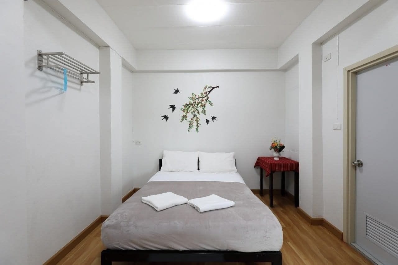 Een keurig opgemaakt bed met een grijze spreidek en twee gevouwen witte handdoeken, een wandrek voor handdoeken, een klein tafeltje met een rode tafelkleed en een vaas met bloemen, en een deur aan de rechterkant.