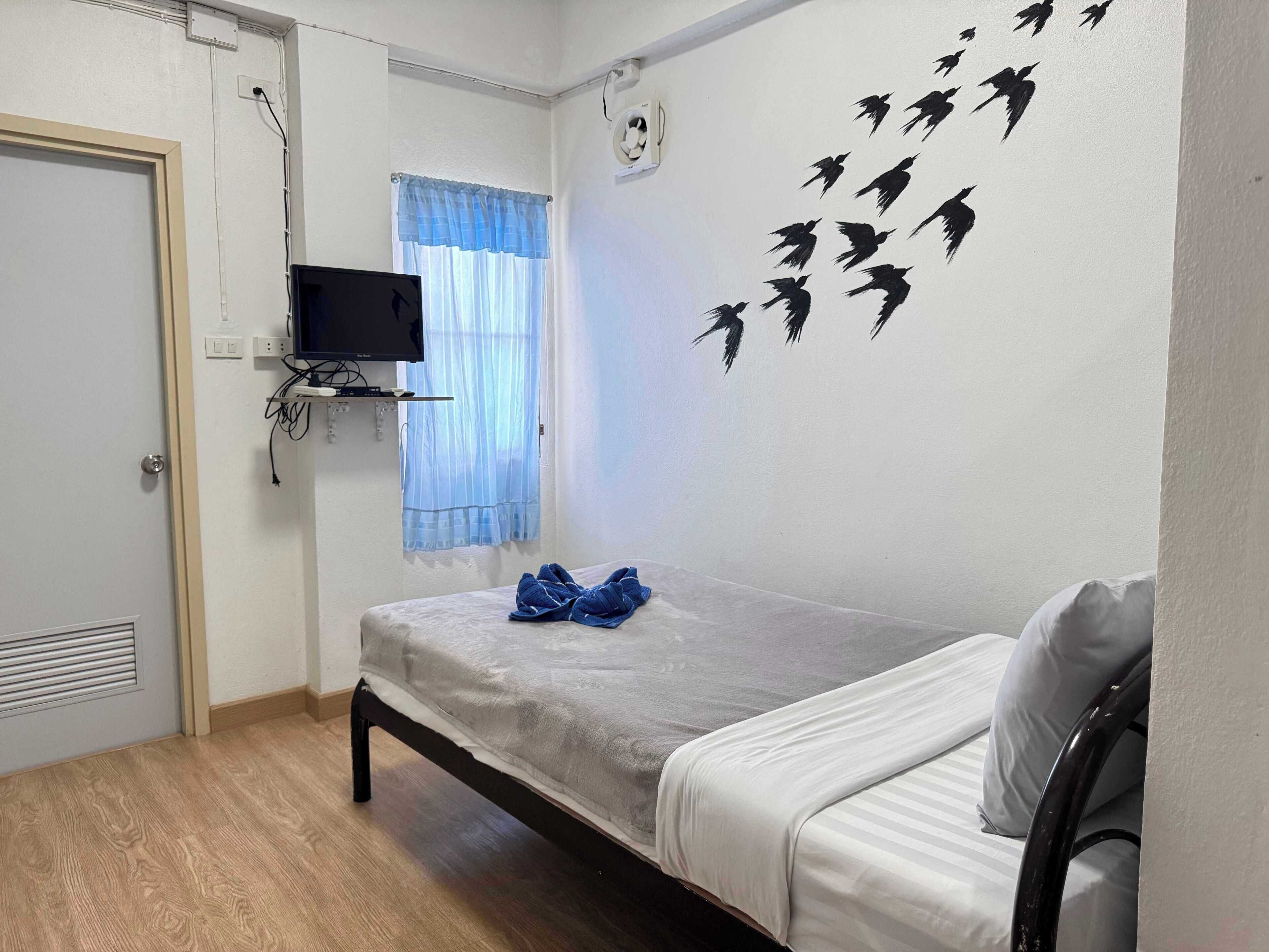 Een kleine, schone kamer met een eenpersoonsbed, een aan de muur bevestigde televisie en een raam met dunne gordijnen.