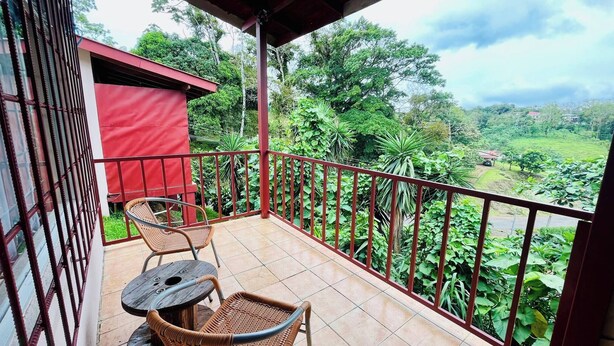 A balcony with wicker furniture, a red building, and a view of a lush green landscape. - Cozy & Beautiful Casa Aire, Near Lake Park & Town (Arenal)