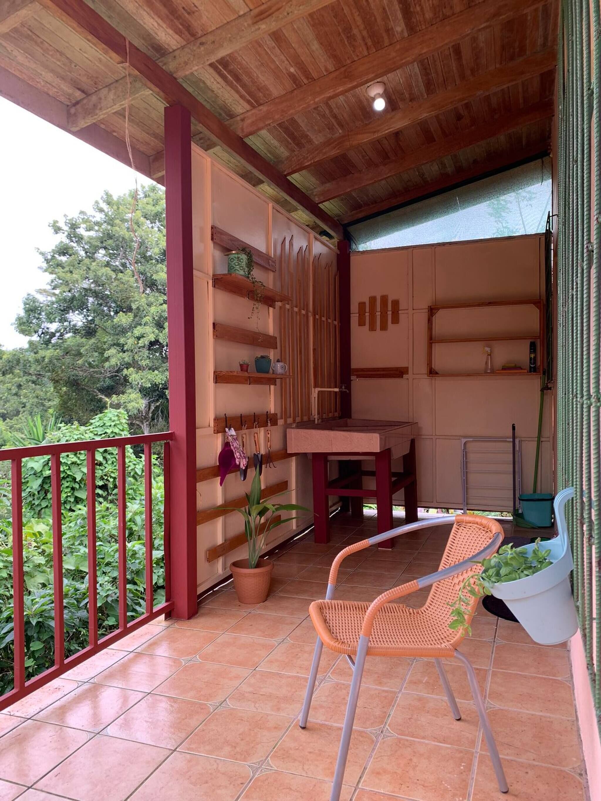 A covered balcony with a wooden ceiling, a table, two wicker chairs, and a potted plant.