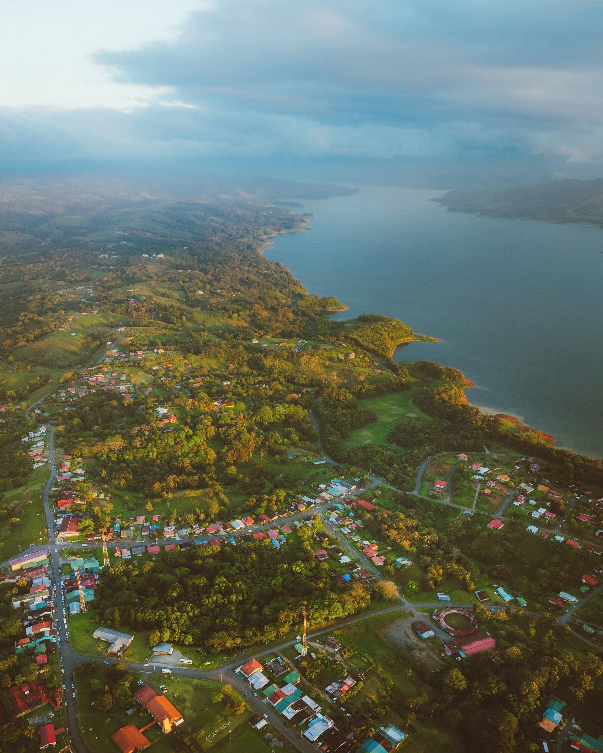 An aerial view of a coastal town with a mix of residential and commercial buildings, a prominent road, and a body of water.