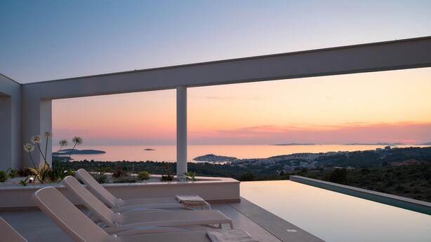 A modern outdoor lounge area with a pool, overlooking a scenic view of a coastal town during sunset.