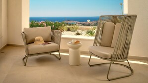 Two wicker chairs with cushions and a small table with a croissant and a cup, overlooking a scenic view of the sea and greenery.