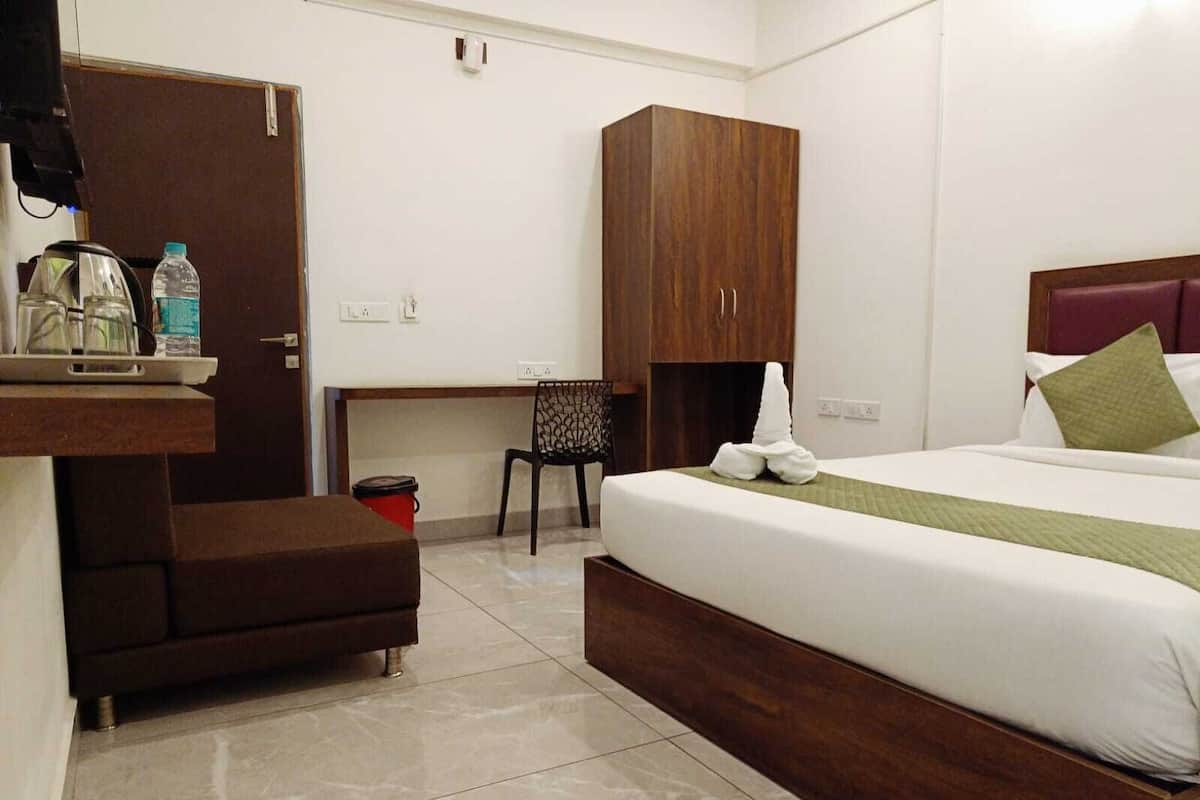 A hotel room with a bed, a wooden wardrobe, a desk with a chair, a small table with a water bottle, and a trash bin.