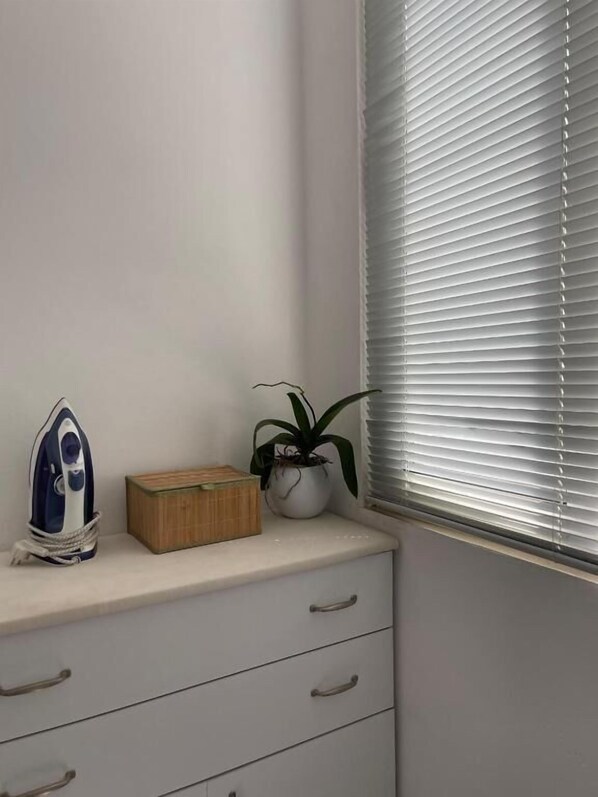 Un coin de pièce avec une commode blanche, une boîte en bambou, une plante en pot et une planche à repasser fixée au mur.