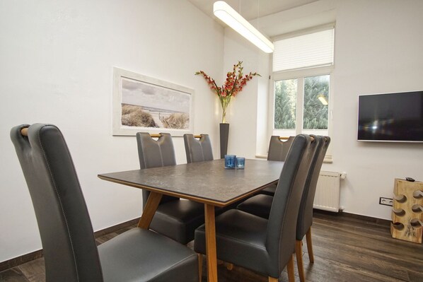 A modern dining room with a rectangular table, six leather chairs, a vase with red flowers, a framed picture on the wall, and a flat-screen TV mounted on the wall.