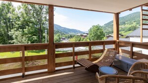 A wooden deck with a view of a mountainous landscape, a lounge area with a table and chairs, and a wooden railing. - L`Altima Megève - Eclat des Veriaz (Megève)