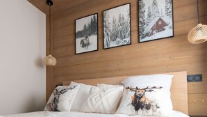 A bedroom with a wooden headboard, a bed with white bedding, and three framed pictures on the wall depicting snowy landscapes and a cabin. - L`Altima Megève - Eclat des Veriaz (Megève)