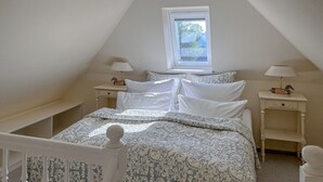 Un dormitorio con techo inclinado, una ventana, una cama con colcha estampada, dos mesitas de noche con lámparas y una mesita auxiliar.