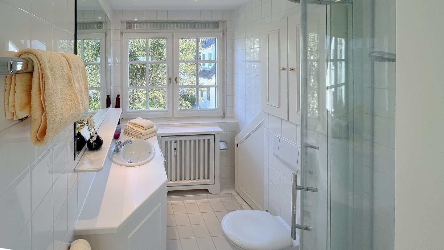 A bathroom with a white sink, toilet, and shower. There is a window with a view of greenery outside.
