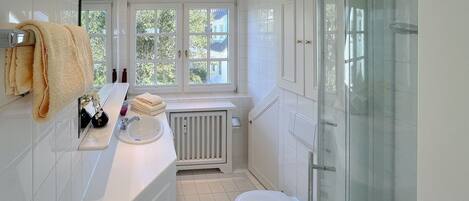 A bathroom with a white sink, toilet, and shower. There is a window with a view of greenery outside.