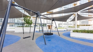 A playground with swings and a blue rubberized surface, surrounded by a building with large windows and balconies. - Meydan Apartment Sleeps 4 With Great Amenities (Dubai)