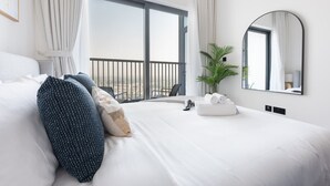 A hotel room with a large bed, white linens, a round mirror, a potted plant, and a view of the outdoors. - Meydan Apartment Sleeps 4 With Great Amenities (Dubai)