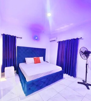 A bedroom with a large bed, blue curtains, a standing fan, and a ceiling light. - Tristone Hotel&Aspartment (Lekki)