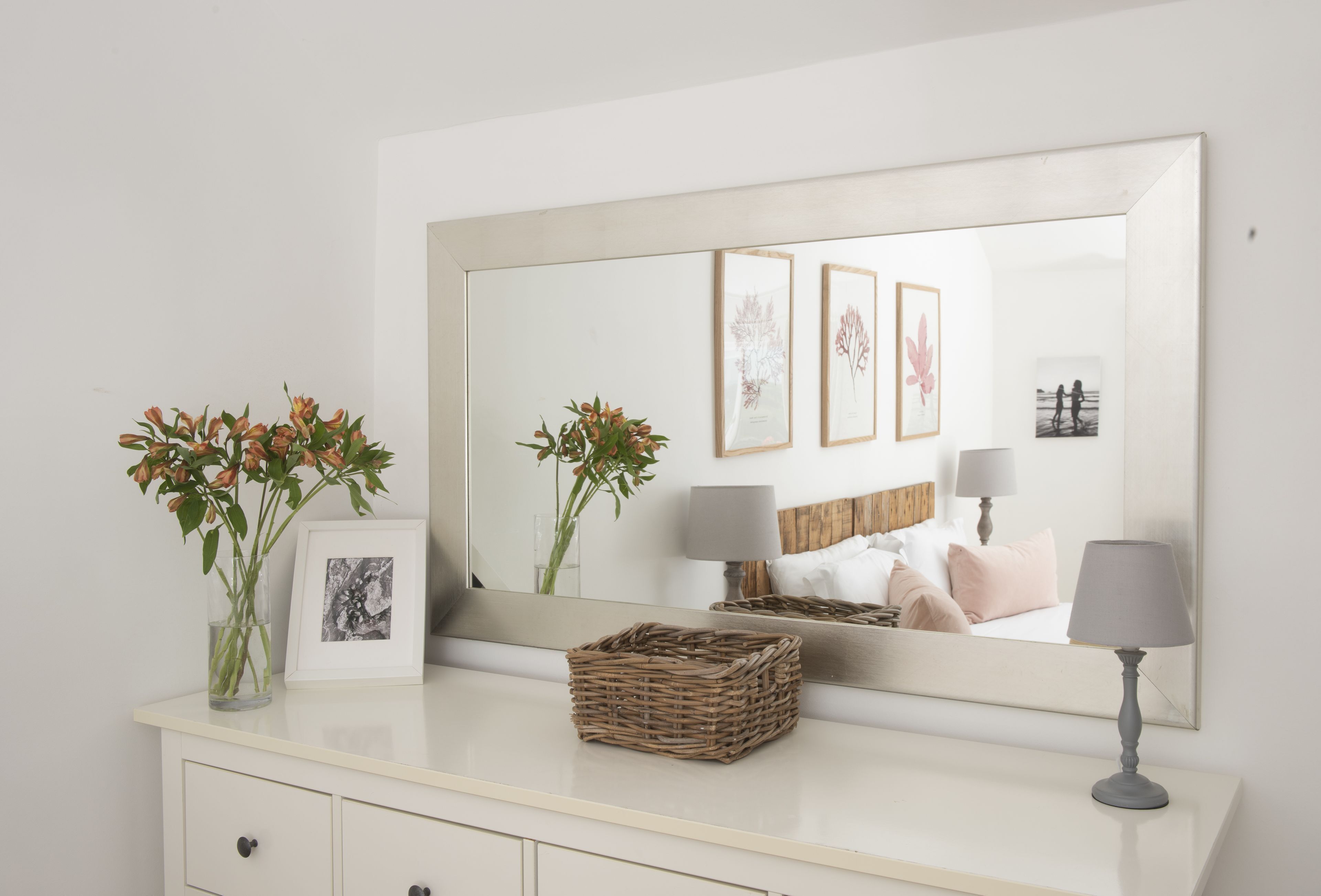 A bedroom with a large mirror, a bedside table with a lamp, a framed picture, and a vase with flowers.
