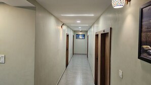 Un pasillo largo y estrecho con paredes de tonos claros y suelo de baldosas. Se observan puertas de madera en un lado y un aplique en la pared en el otro.