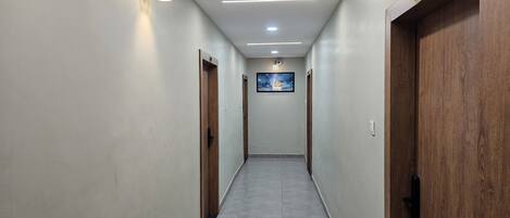 Un pasillo largo y estrecho con puertas de madera a ambos lados, suelo de baldosas e iluminación empotrada en el techo.