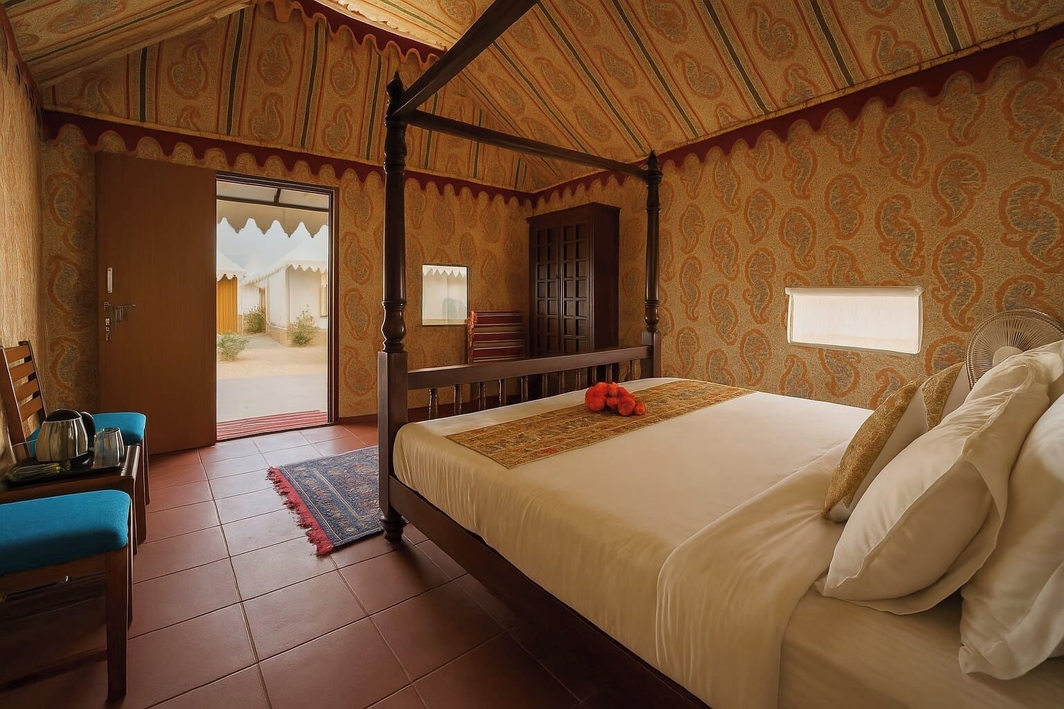 A room with a four-poster bed, a wooden desk with a chair, and a view of a courtyard through an open door.