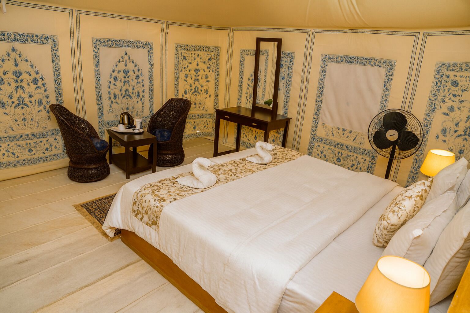 A room with a bed, two chairs, a table, a mirror, a fan, and decorative pillows.