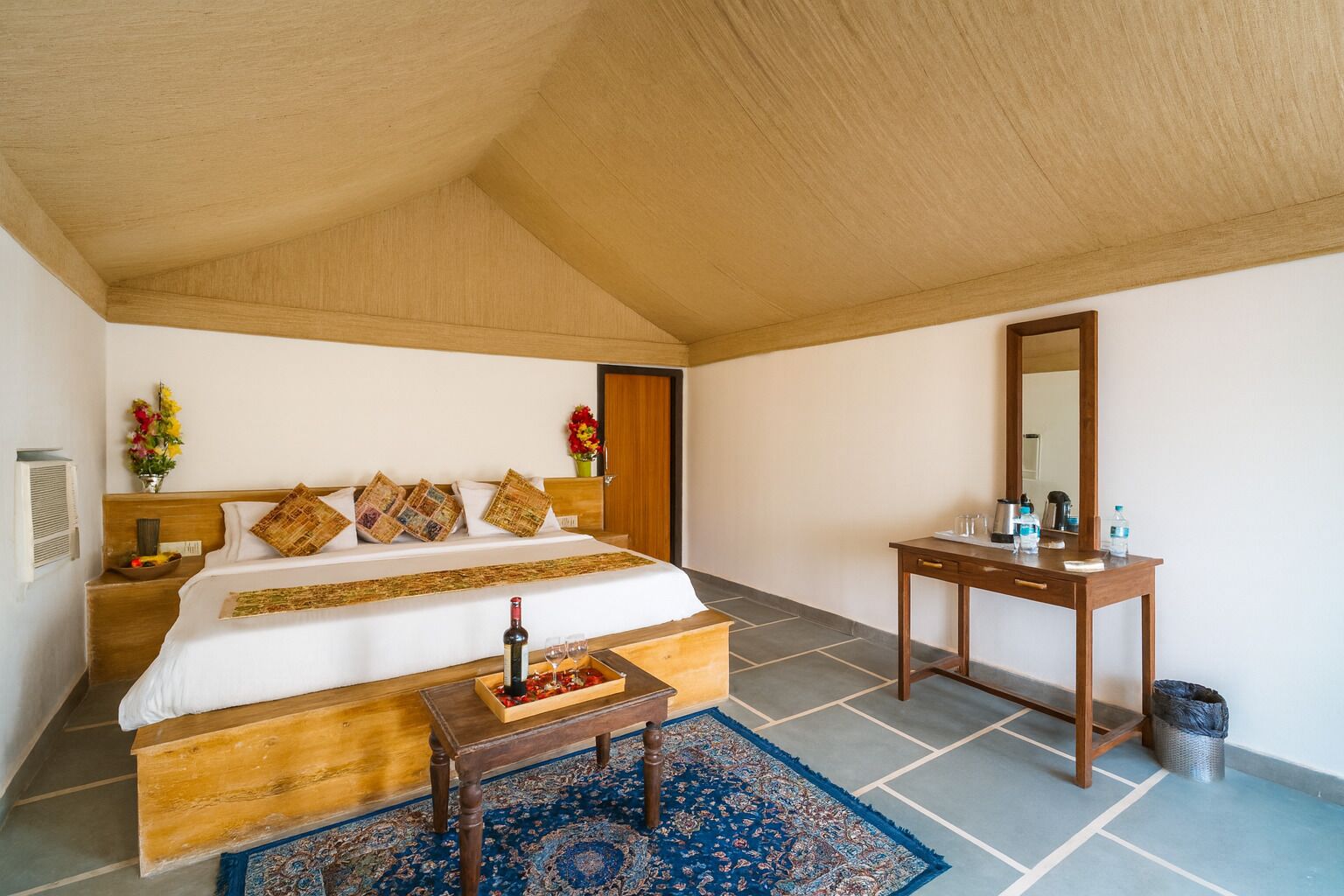 A bedroom with a large bed, a wooden nightstand, a bedside table with a bottle and glasses, a mirror, and a blue patterned rug.
