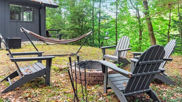 A rustic outdoor seating area with Adirondack chairs and a hammock near a brick fire pit, surrounded by a forested landscape. - Stylish Bungalow near Saugatuck (Fennville)