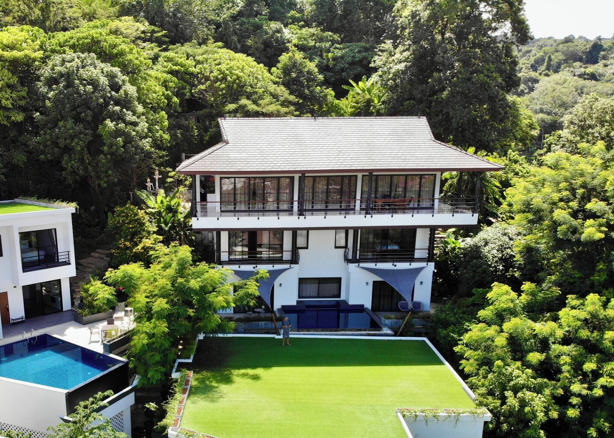 A modern two-story house with a large balcony, surrounded by lush greenery.