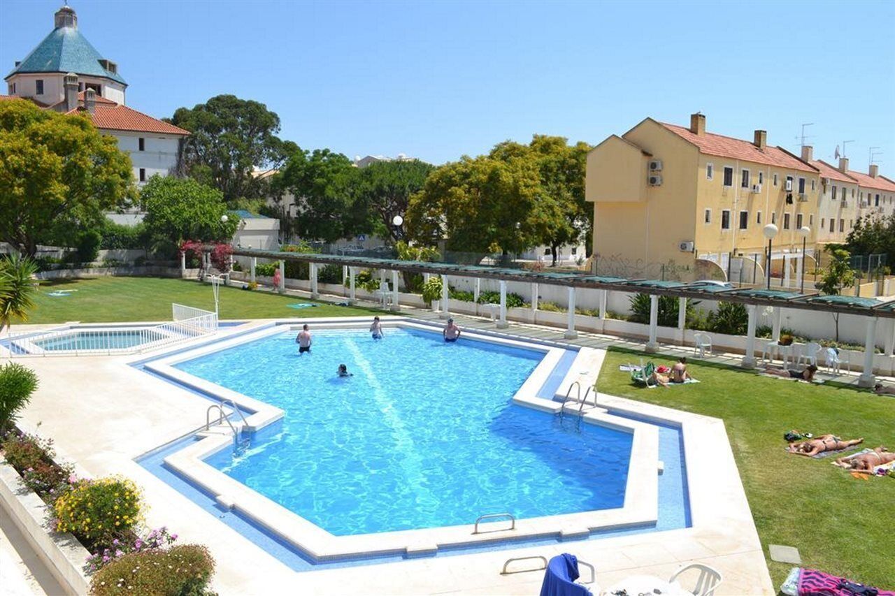 Una piscina con un deck perimetral, un área de descanso techada y vista a edificios al fondo.