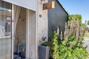 Ein Gebäudeeingang mit einem Schild, auf dem 'Le petit des Ecreviels' steht, einer Nummer '4', einer Tür mit Glasfenster, einem kleinen Hocker und einem Blumentopf mit roten Blüten.