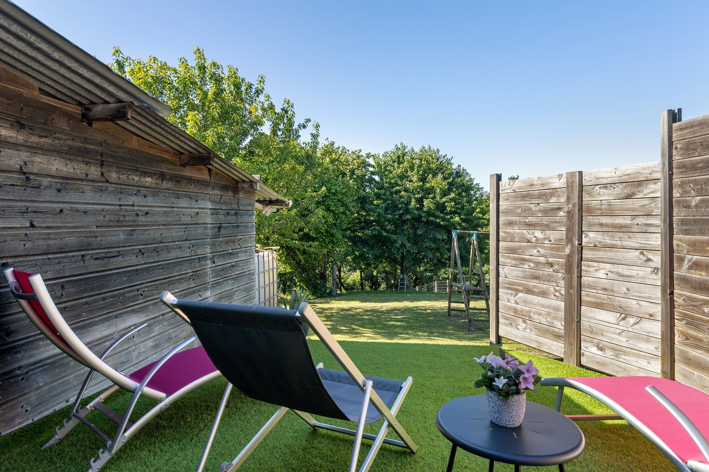 A backyard with a wooden fence, two lawn chairs, a small table with a potted plant, and a swing set.