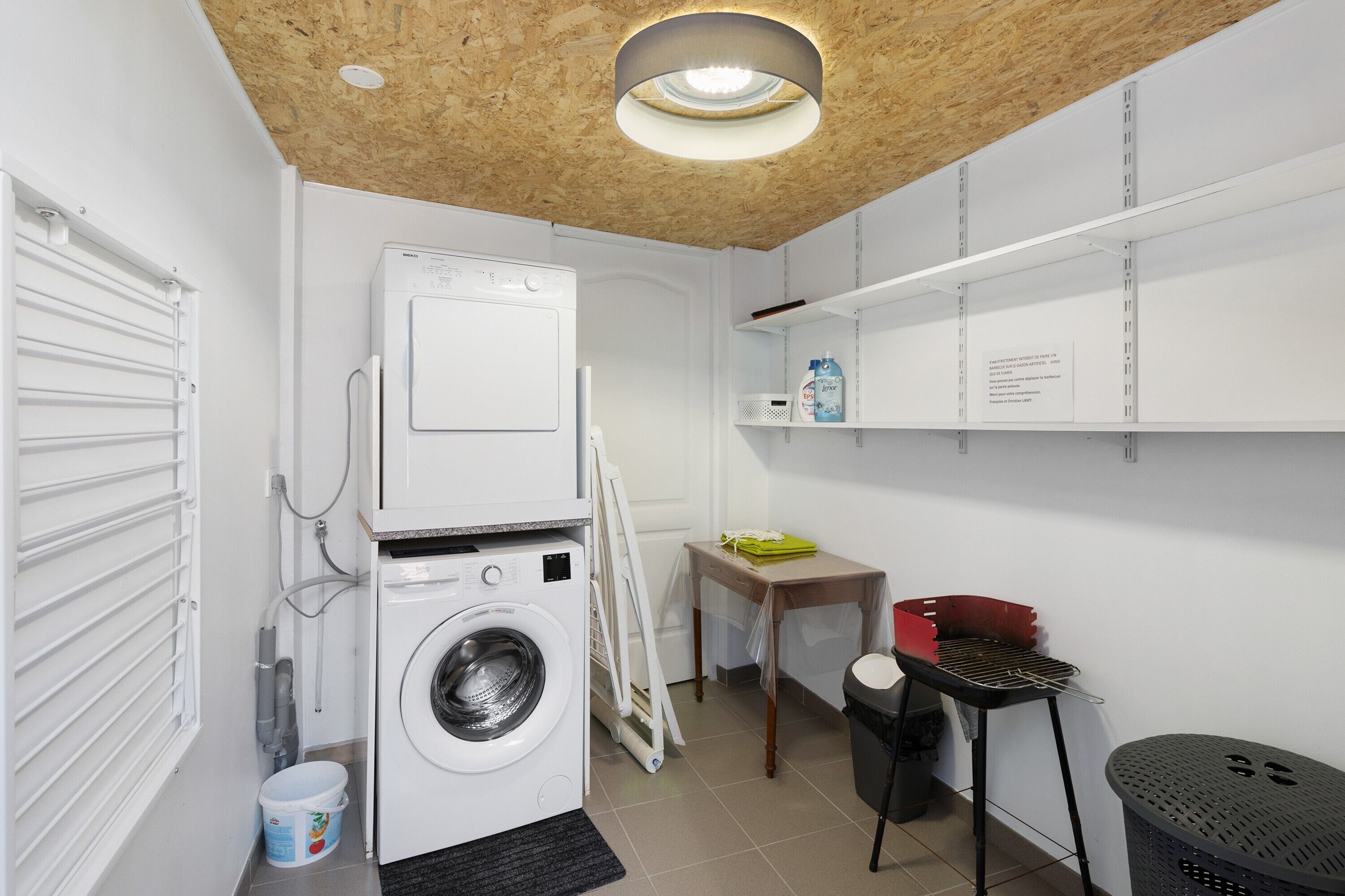 A laundry room with a washing machine, a drying rack, a table with a basket, and a trash bin.