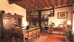 En traditionel spisestue med et træbord, stole og en lysekrone. Der er en trappe til en etage ovenover, og en pejs ses i baggrunden.
