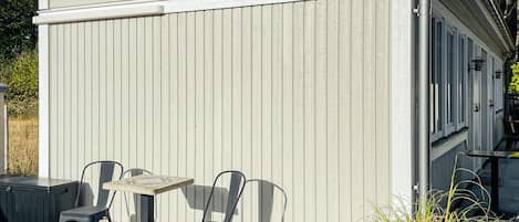 A building with a covered patio area, featuring a table and chairs, a small black object on the ground, and a plant to the right.