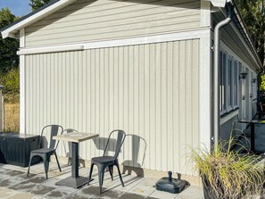 A building with a covered patio area, featuring a table and chairs, a small black object on the ground, and a plant to the right. - 4 Star Holiday Home in Solvesborg (Sölvesborg)
