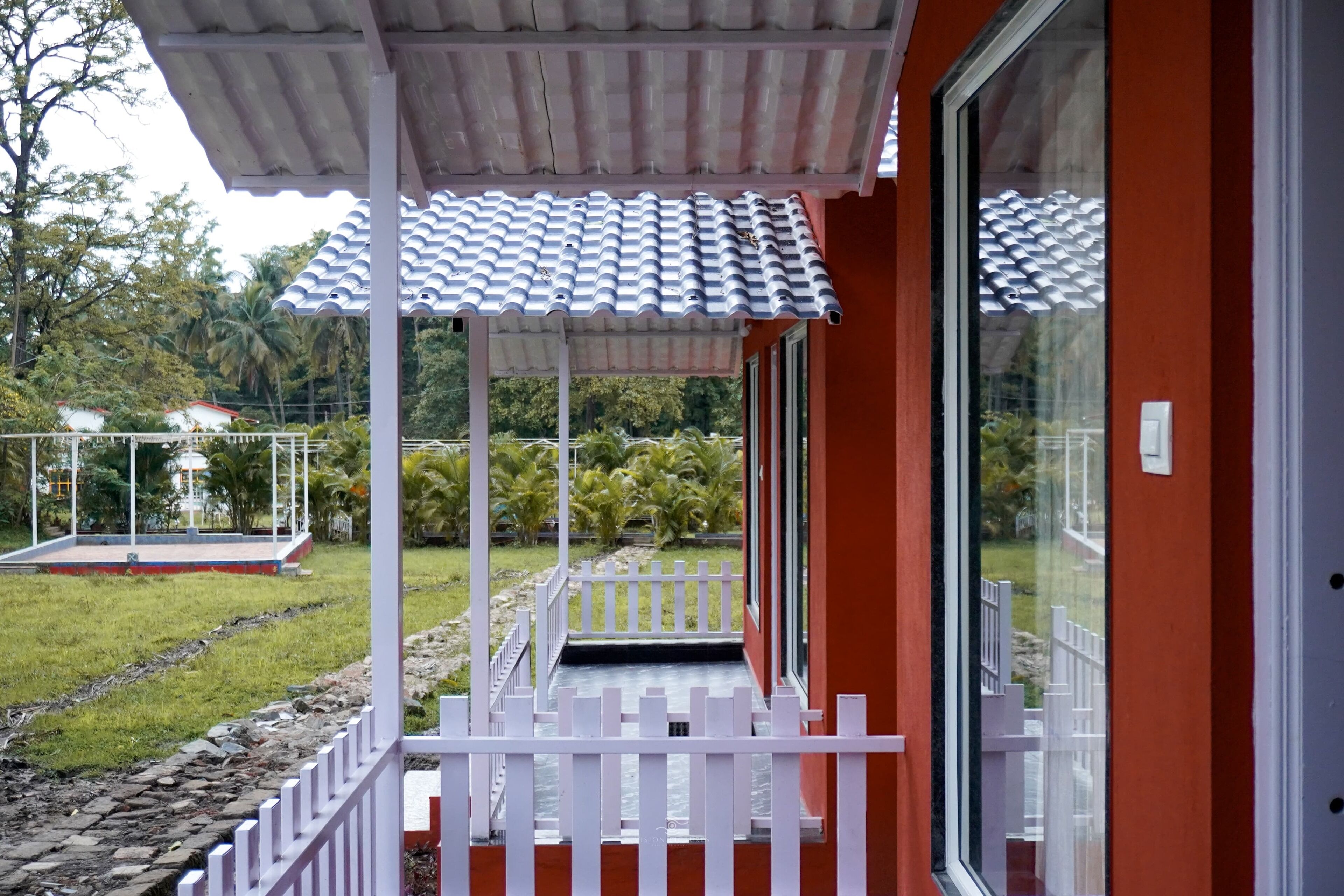 Un espacio al aire libre techado con cerca blanca, techo de baldosas y vista a un jardín con palmeras.