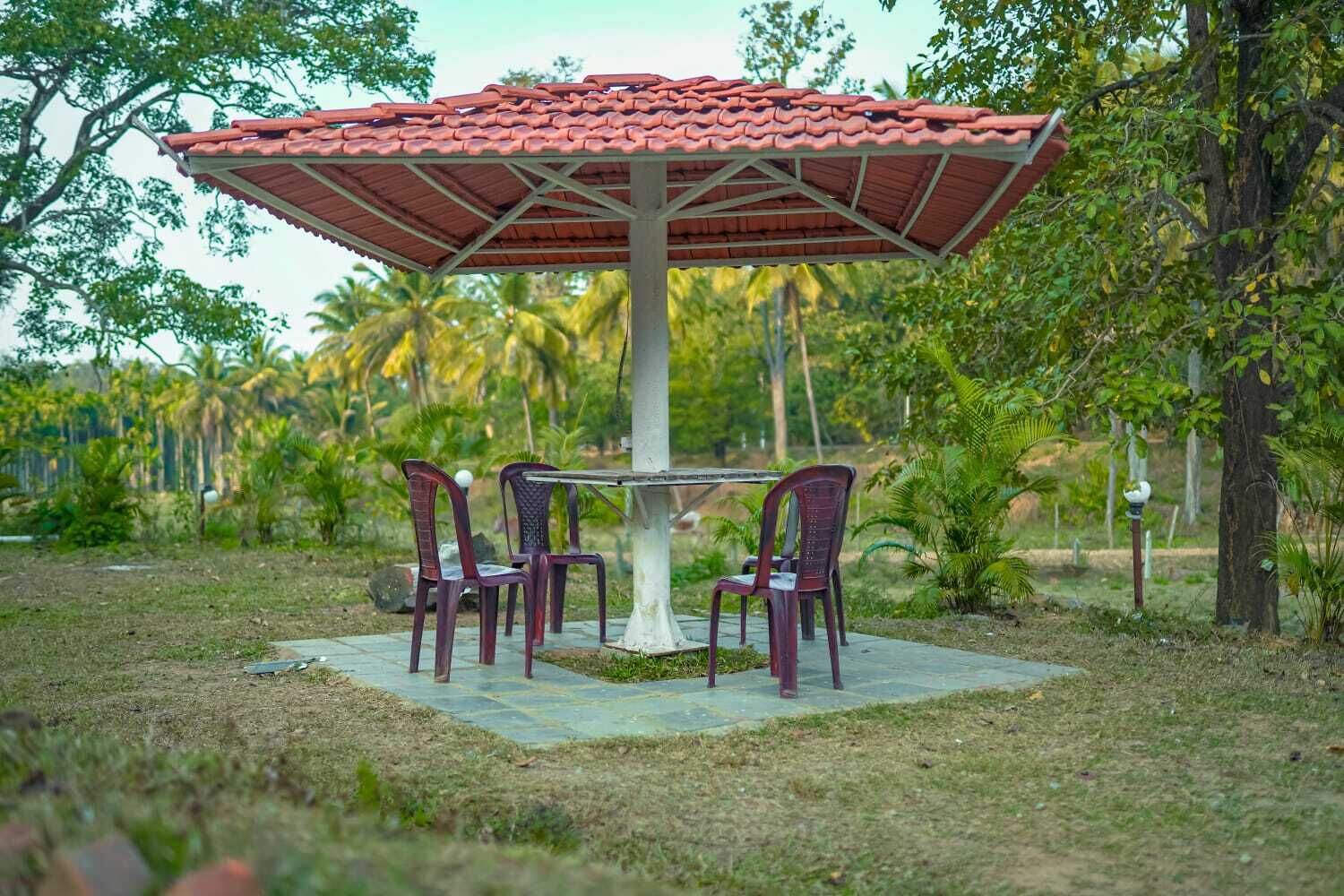 A gazebo with a tiled roof, surrounded by chairs and a table, set in a garden with trees and grass.