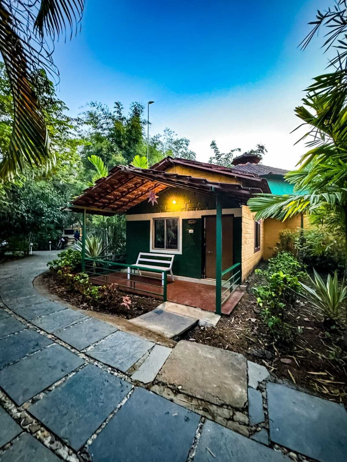 A small, single-story building with a tiled roof and a green door, surrounded by lush greenery and a stone pathway.