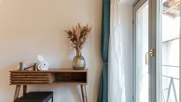 Une pièce avec un bureau en bois, un vase contenant des fleurs séchées, une serviette enroulée et un tabouret noir.