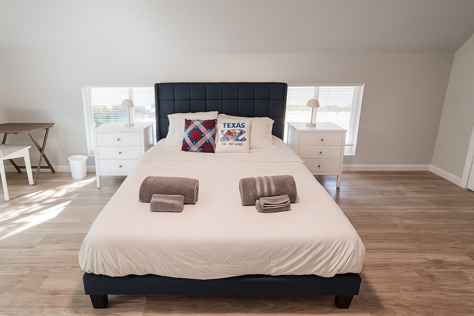 Una cama bien hecha con un cabecero oscuro, dos almohadas y dos toallas dobladas. Hay dos mesitas de noche con lámparas, una mesita pequeña y una silla en la habitación.