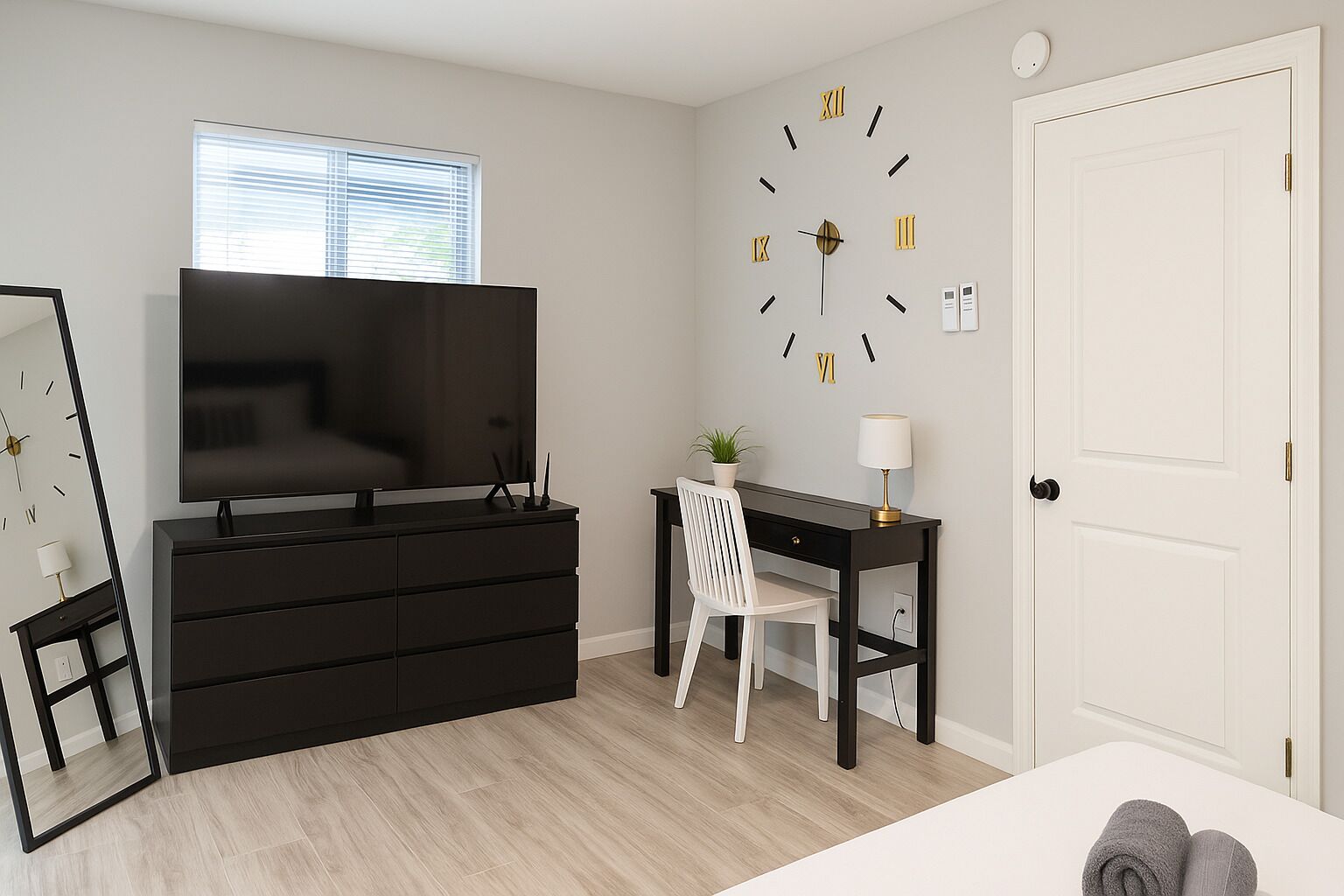 Un dormitorio con una televisión grande, un escritorio con una silla, un reloj de pared y una puerta.