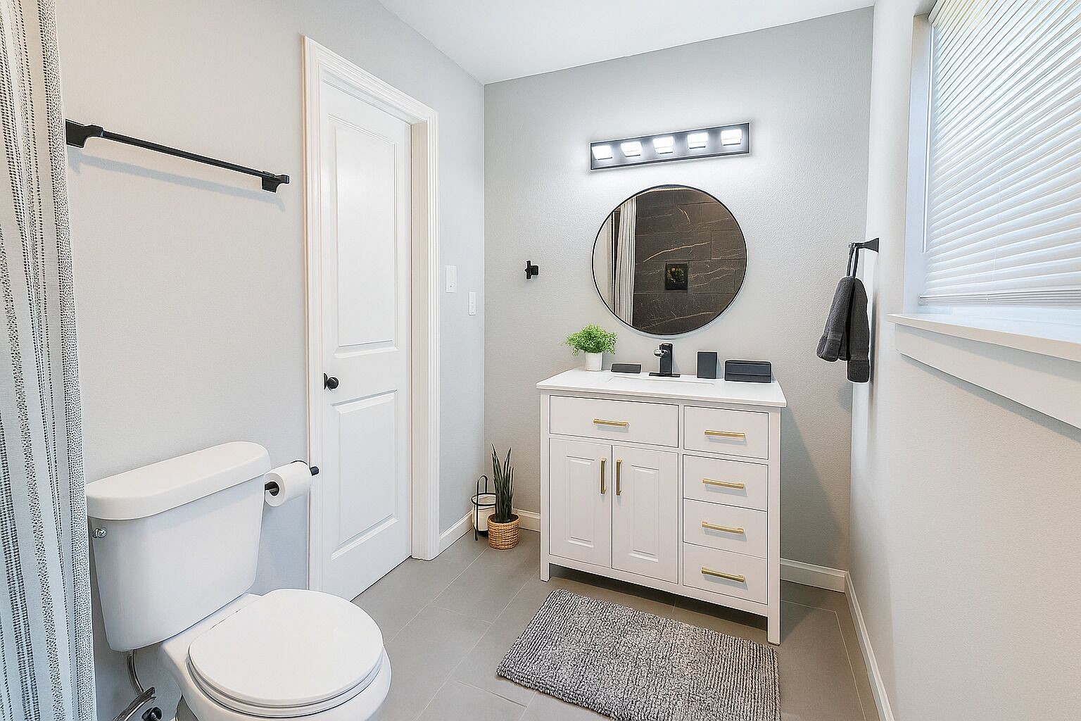 Ein Badezimmer mit Toilette, Waschbecken mit Spiegel, Schubladenschrank, kleiner Pflanze und Fenster mit Jalousien.