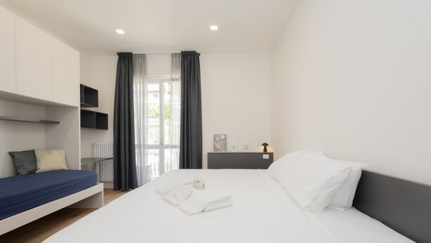 A modern hotel room with a large bed, a smaller bed, a desk, a chair, a TV, and a window with curtains. - Milano Centro | Fontana House  (Milano)